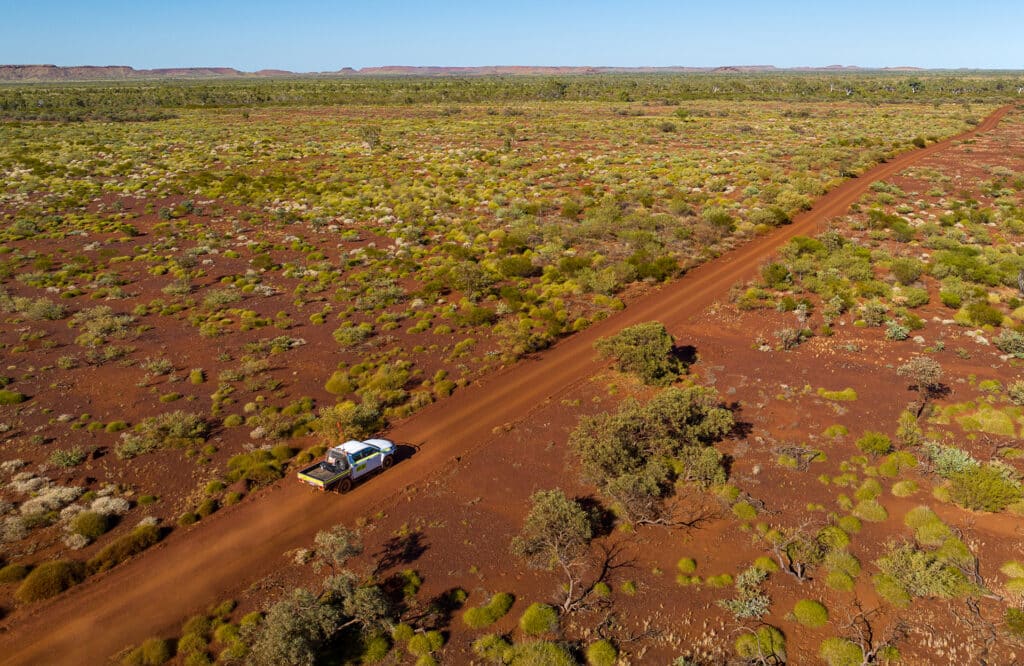 Aerial view of a long dirt road cutting through remote Australian bushland, with a work vehicle driving along.