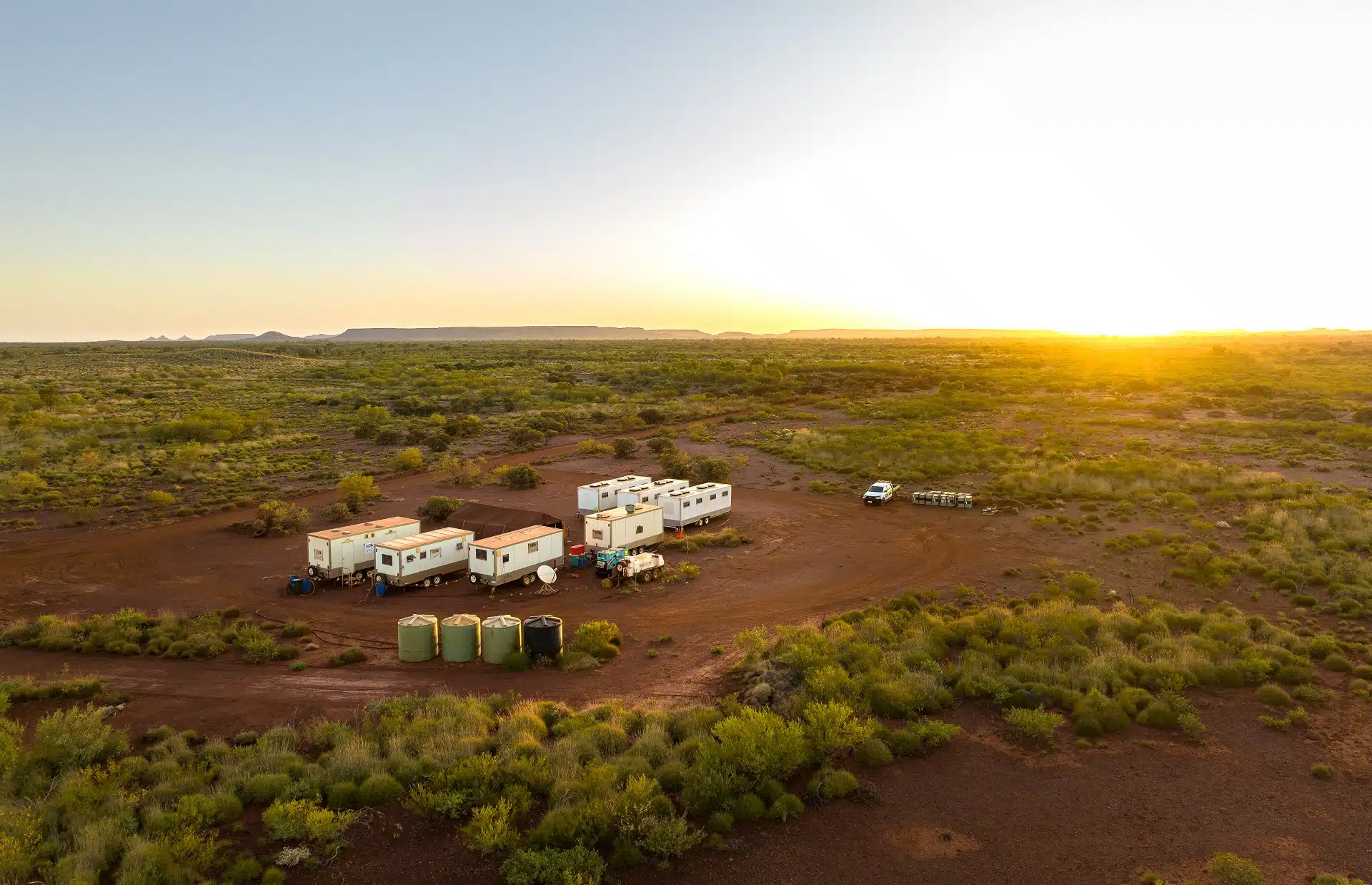 Aerial view of a remote work camp with portable buildings, water tanks, and a dirt access road, set against a sunset landscape.