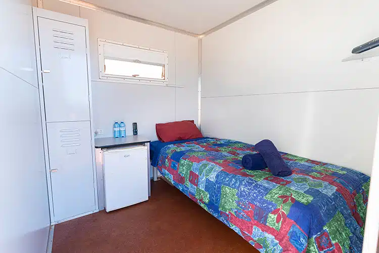 Compact accommodation room with a single bed, storage locker, mini-fridge, and neatly arranged bedding in a portable unit.