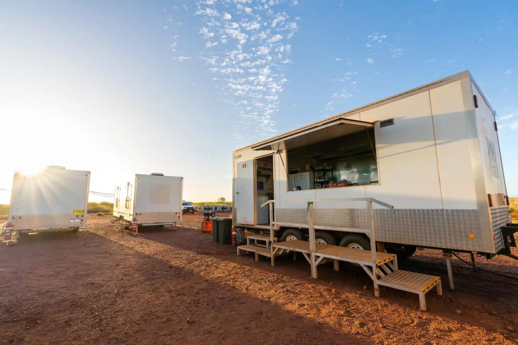 Mobile kitchen trailer with open serving window and stairs, set up at a remote worksite under a clear morning sky.